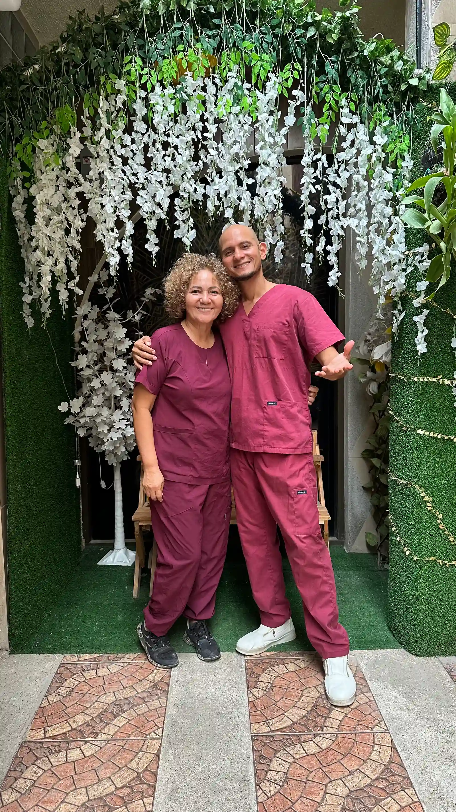 Dos integrantes de SIRONA posan con uniforme color vino bajo un arco de plantas y flores blancas.
